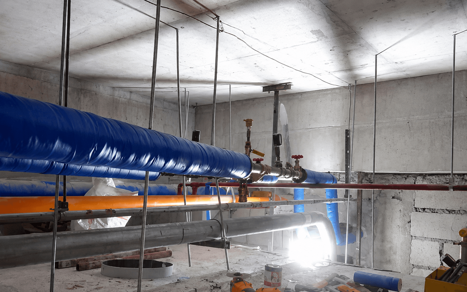 Insulated blue and orange pipes hanging from a concrete ceiling in an industrial basement.