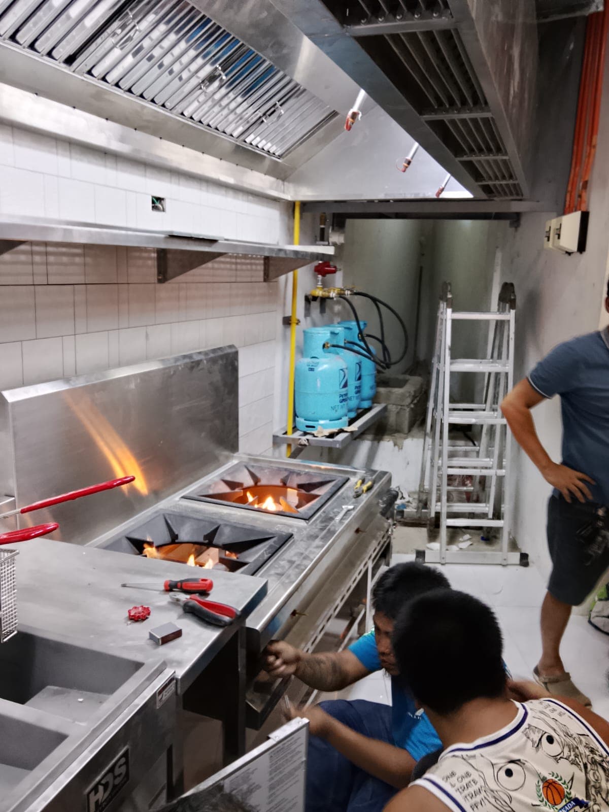 Technicians working on a commercial gas stove with lit burners in a professional kitchen.