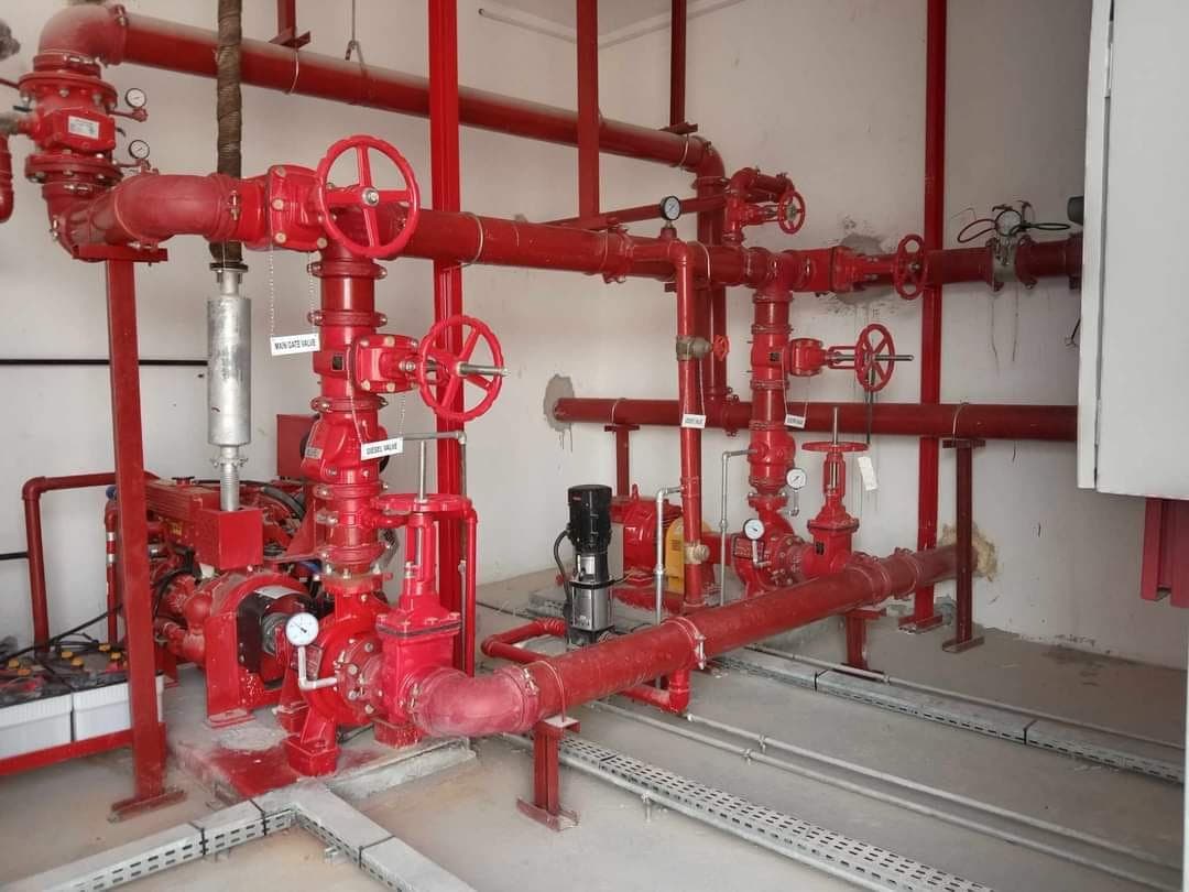 Industrial fire pump system featuring red pipes, large handwheel valves, and pressure monitoring gauges.